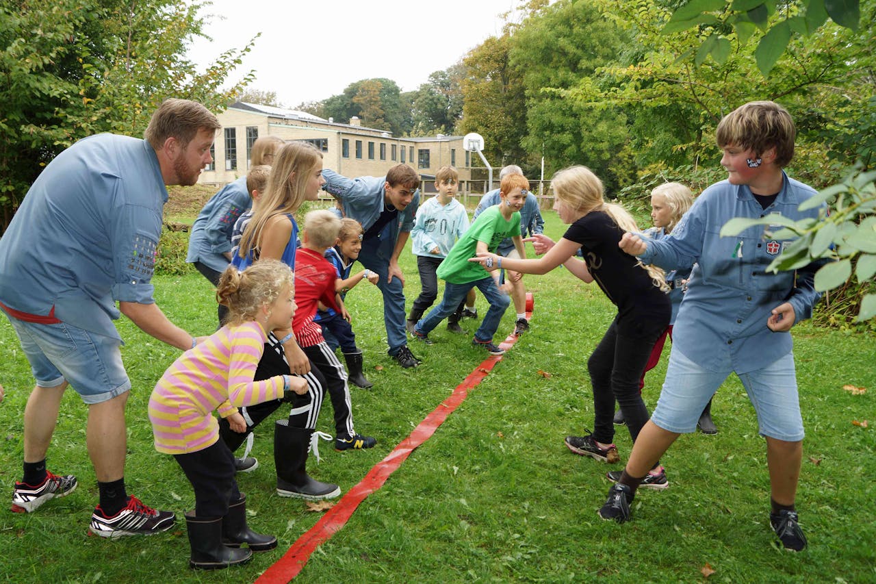 Een groep kinderen speelt buiten een spelletje. Op de grond ligt een rode scheidingslijn.