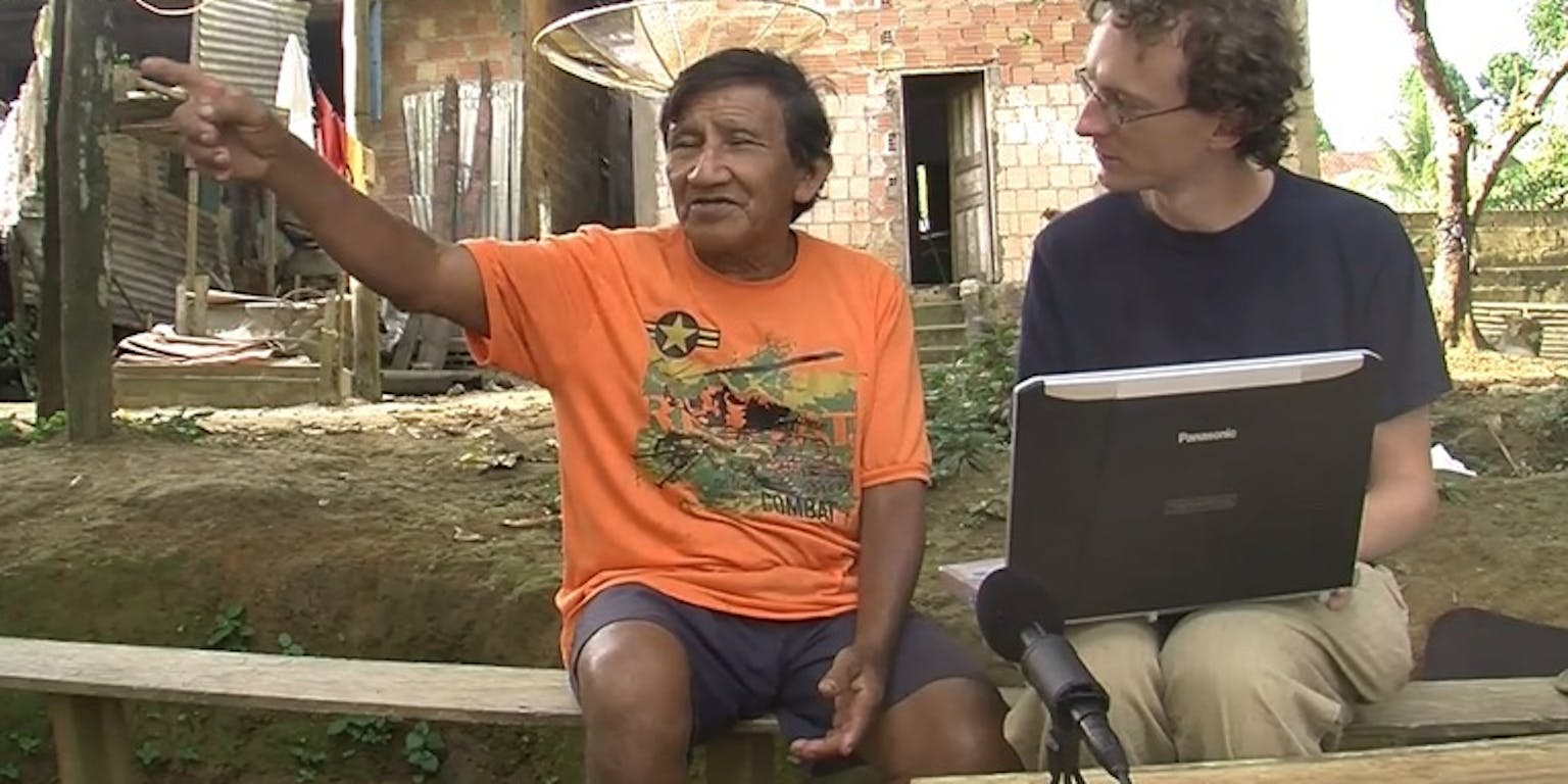 Twee mannen zitten op een bankje met een laptop en een microfoon.