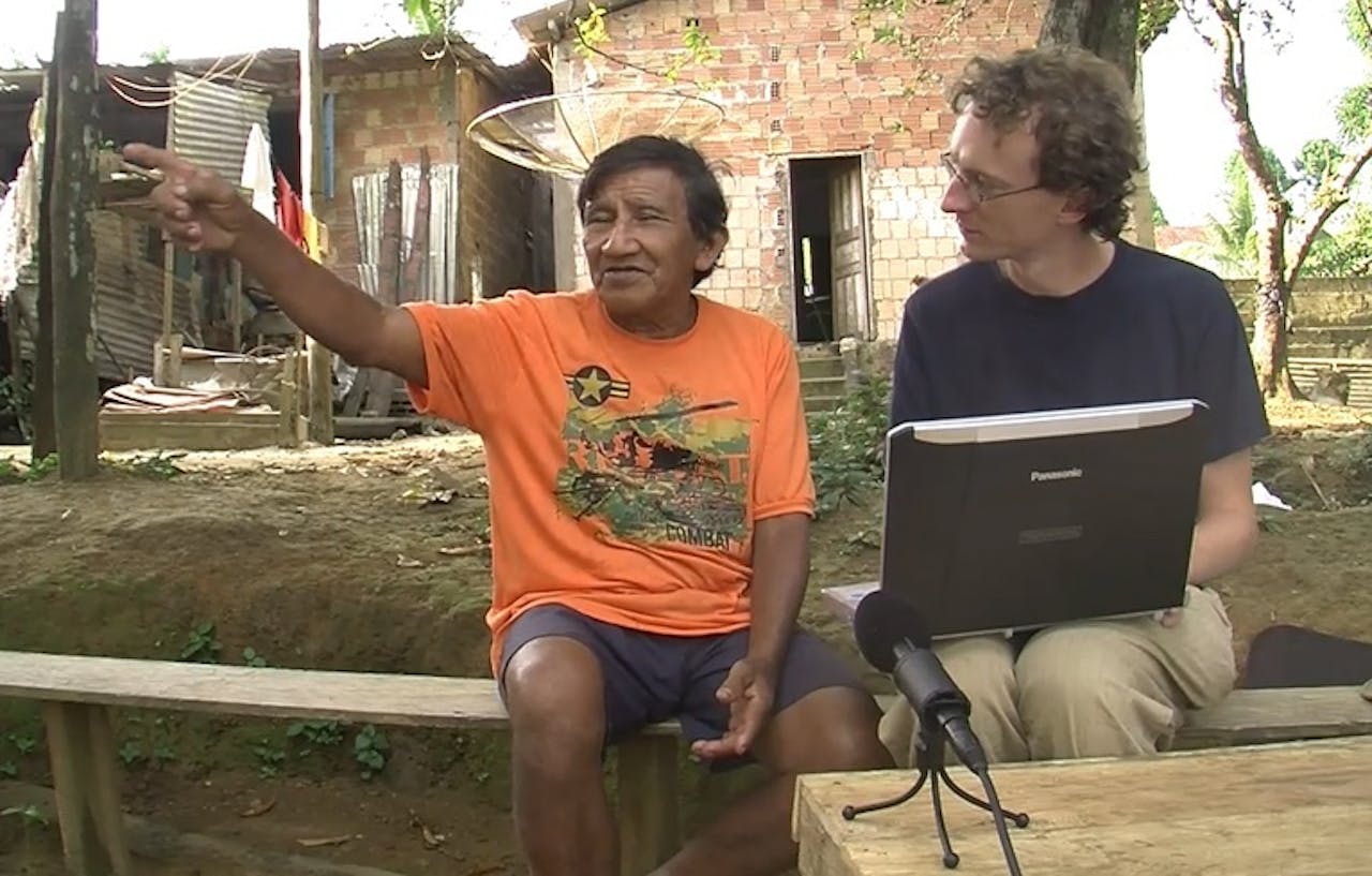 Twee mannen zitten op een bankje met een laptop en een microfoon.