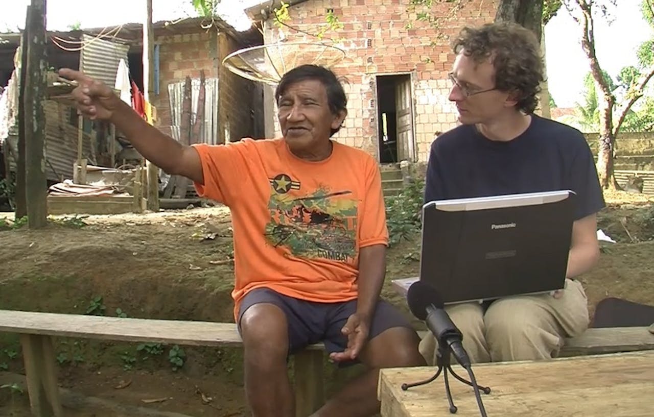Taalonderzoeker Simeon Floyd in gesprek met een man die Nheengatú spreekt. De man wijst naar de stand van de zon om vier uur in de middag. Hij heeft in zijn taal geen woorden om hetzelfde uit te drukken. Twee mannen zitten op een bankje met een laptop en een microfoon.