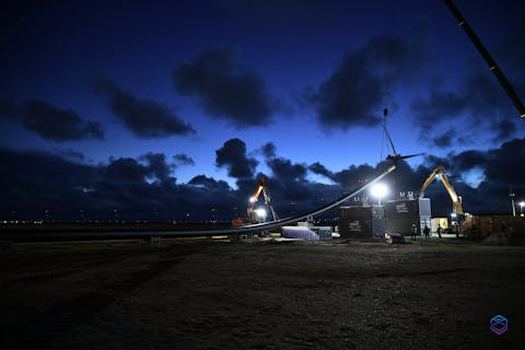 Aanleg van de CO2-leiding op de Maasvlakte. Een lange zwarte pijpleiding loopt over de grond en wordt door twee kranen in de lucht getild. Een grote bouwlamp verlicht het werkgebied. De lucht en omgeving zijn donker.