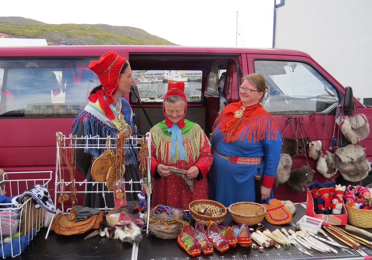 Een foto van drie lappen. Deze bevolkingsgroep draagt klederdracht en staat voor een rood busje waren te verkopen op een markt.