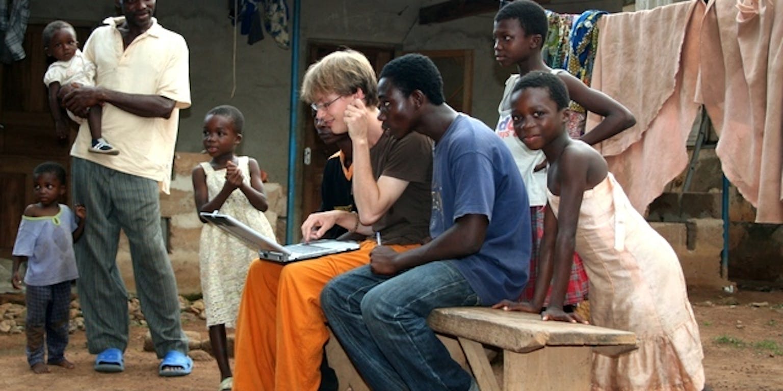 Drie volwassenen en vijf kinderen staan rondom of zitten op een bankje. Een persoon heeft een laptop op schoot.