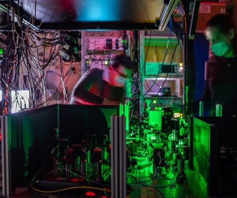 Twee onderzoekers met mondkapje staat bij een groen verlicht knooppunt van een quantumnetwerk. Dat bestaat uit veel kabels, spiegels en filters, die laserstralen naar een diamantchip leiden.