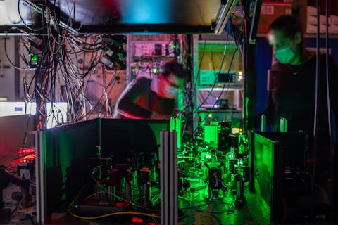 Sophie Hermans en een collega aan het werk bij een van de quantumnetwerk-knooppunten bij onderzoeksinstituut QuTech in Delft. Twee onderzoekers met mondkapje staat bij een groen verlicht knooppunt van een quantumnetwerk. Dat bestaat uit veel kabels, spiegels en filters, die laserstralen naar een diamantchip leiden.