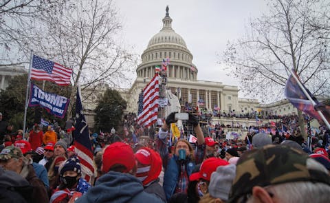 De menigte buiten tijdens de bestorming van het Amerikaanse Capitool op 6 januari 2021. Een menigte met Amerikaanse vlaggen en Trump-spandoeken voor en op de trappen van het Capitool, een wit gebouw met zuilen en koepel.