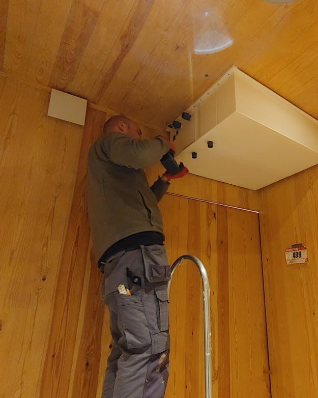 Een man in grijze werkkleding staat op een ladder in een ruimte met houten wanden en een houten plafond. Tegen dat plafond bevestigt hij een witte vierkanten tank. Een man in grijze werkkleding staat op een ladder in een ruimte met houten wanden en een houten plafond. Tegen dat plafond bevestigt hij een witte vierkanten tank.