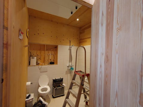 Installatie van het Regenwatertoilet in The Green Village bij de TU Delft. Een toiletruimte met witte tegels en houten wanden en plafonds. In de ruimte staan een ladder en een gereedschapskist.
