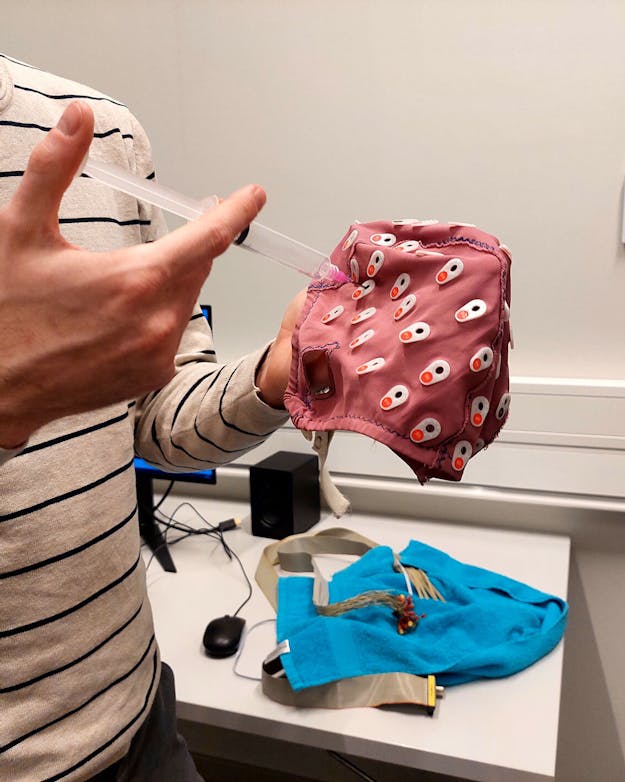De welbekende EEG-muts. In de spuit zit een geleidende gel die de elektroden in de muts helpt de zwakke hersensignalen op te pikken. Onderzoeker met een rode EEG-muts en een spuit met geleidende gel in zijn handen.