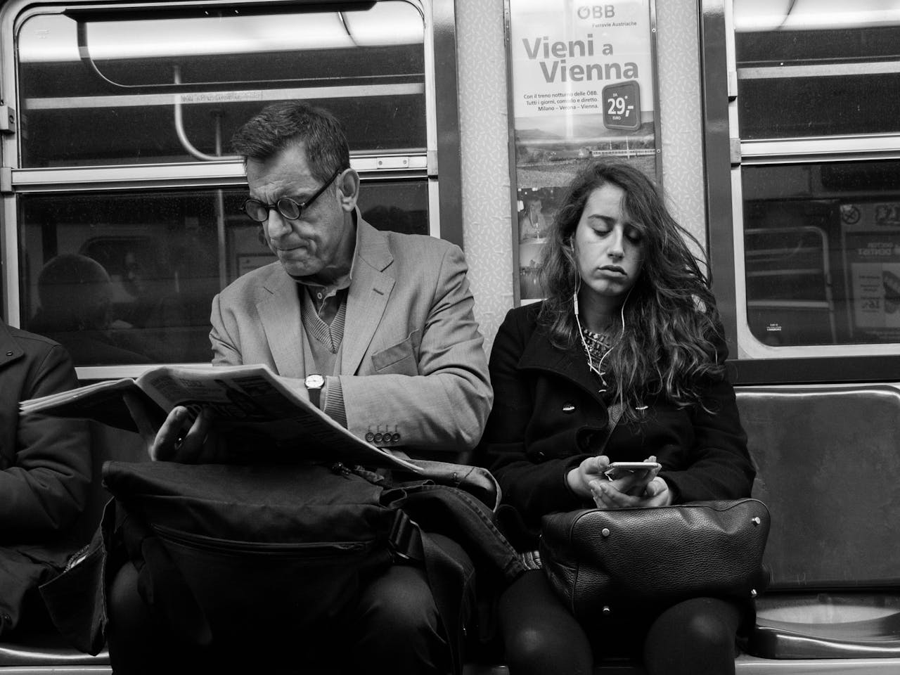 Twee personen in de metro. Een persoon leest de krant, de andere persoon zit op een smartphone en heeft oortjes in.