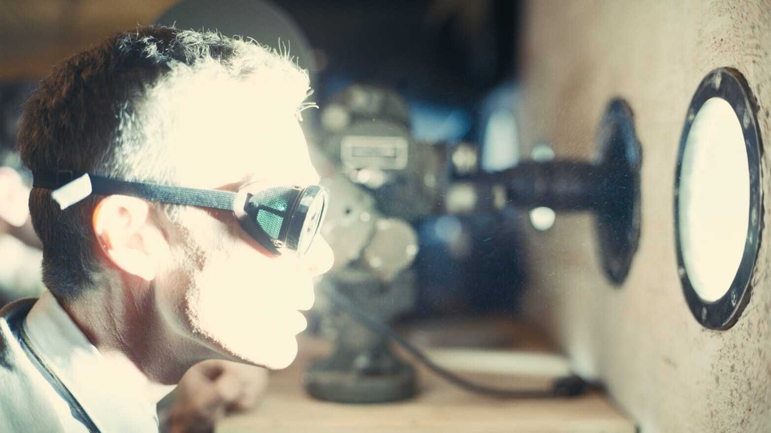 Een man kijkt vanuit een bunker door een vreemd ogende bril naar een enorm fel licht, veroorzaakt door de ontploffing van een atoombom. Achter hem staat apparatuur.