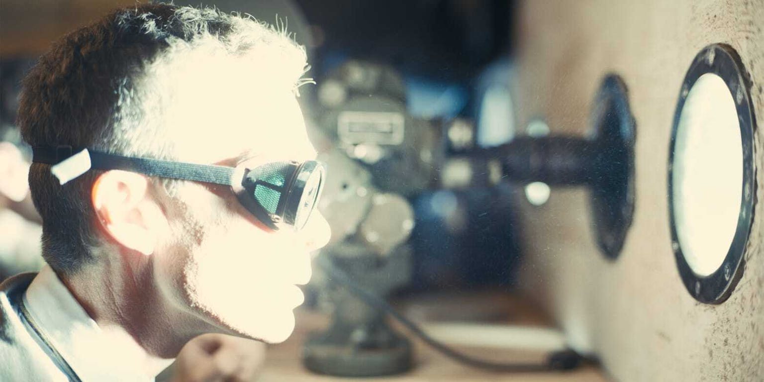 Een man kijkt vanuit een bunker door een vreemd ogende bril naar een enorm fel licht, veroorzaakt door de ontploffing van een atoombom. Achter hem staat apparatuur.