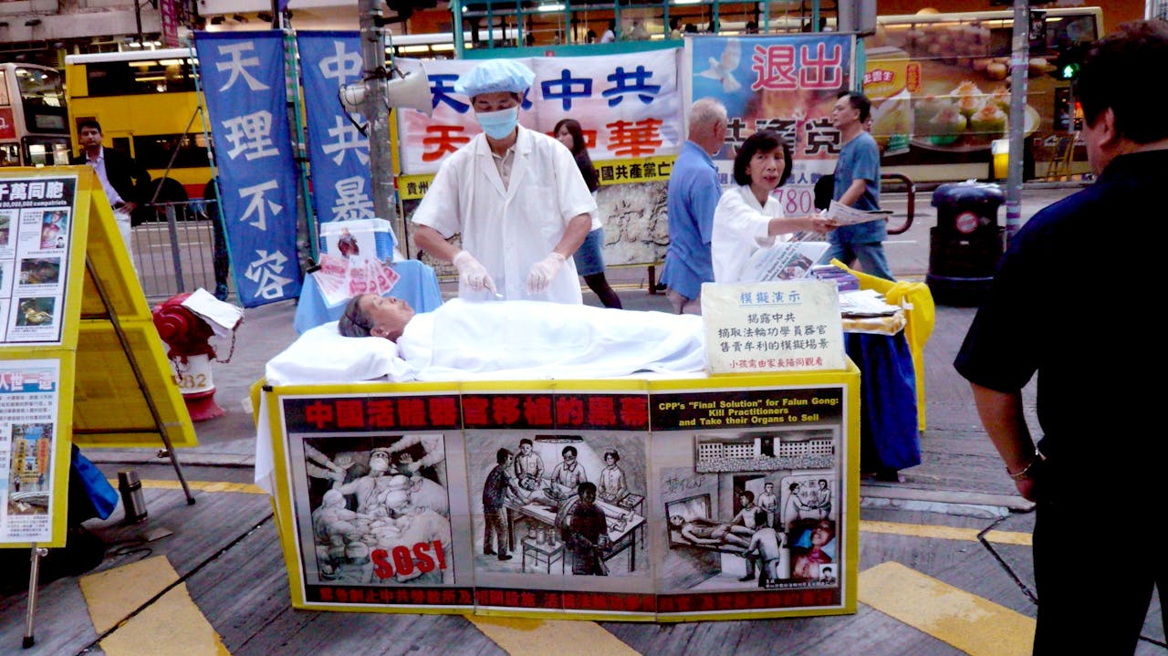 Twee mensen die een rollenspel van dokter en (overleden) patient uitbeelden op een drukke straat in China. Het is een vorm van protest tegen de praktijken van orgaanhandel in China.