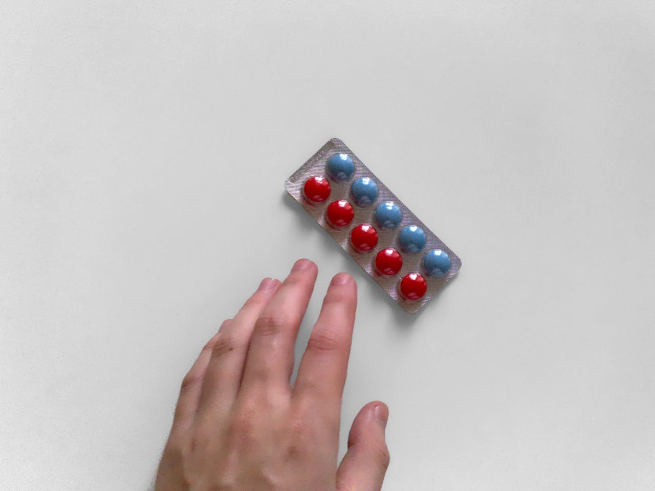 Een pillenstrip met blauwe en rode pillen. Een hand reikt naar de pillenstrip. De achtergrond is wit.