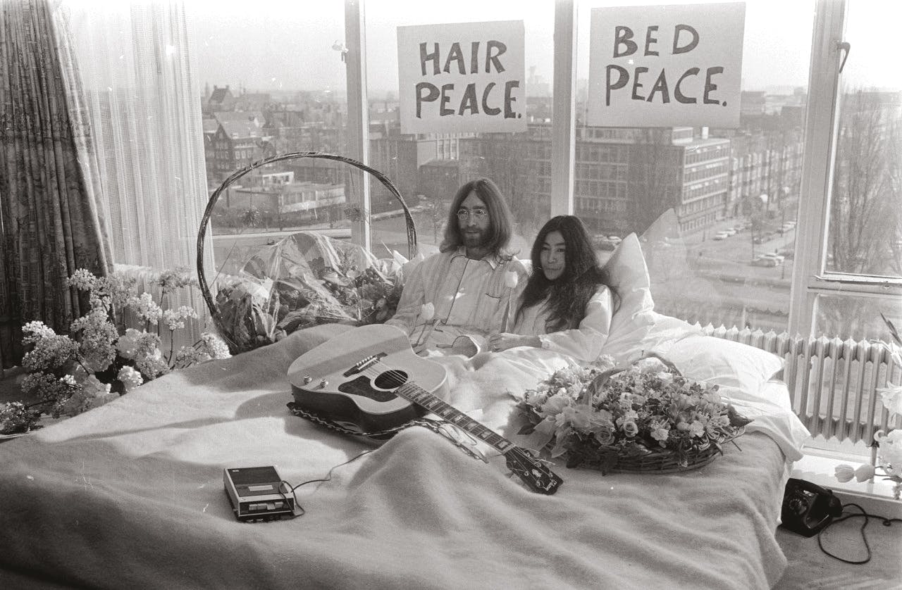 Een zwart-wit foto van John Lennon en zijn echtgenote Yoko Ono in bed met tulpen en een gitaar.
