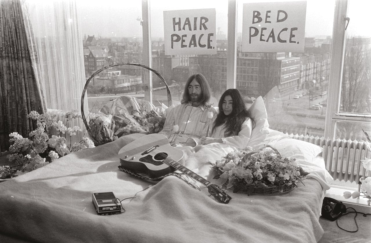 John Lennon zag meer in ‘Hair & Bed Peace’ dan in religie. Een zwart-wit foto van John Lennon en zijn echtgenote Yoko Ono in bed met tulpen en een gitaar.