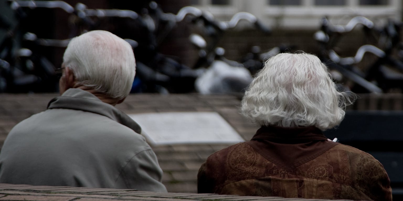 Een ouder echtpaar zit samen op een bankje. De foto is vanaf de achterzijde genomen.