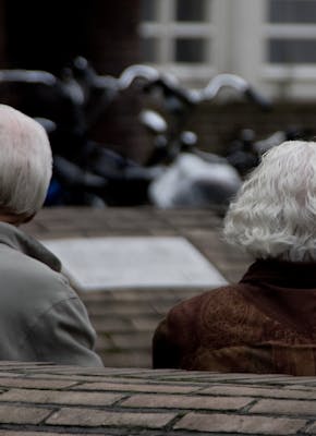 Een ouder echtpaar zit samen op een bankje. De foto is vanaf de achterzijde genomen.