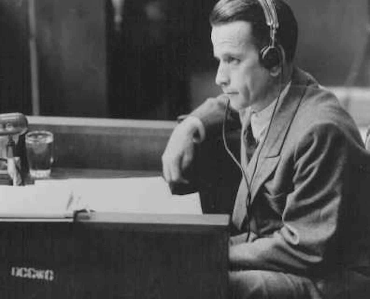 Een man in een pak zit aan een bureau met een koptelefoon op.