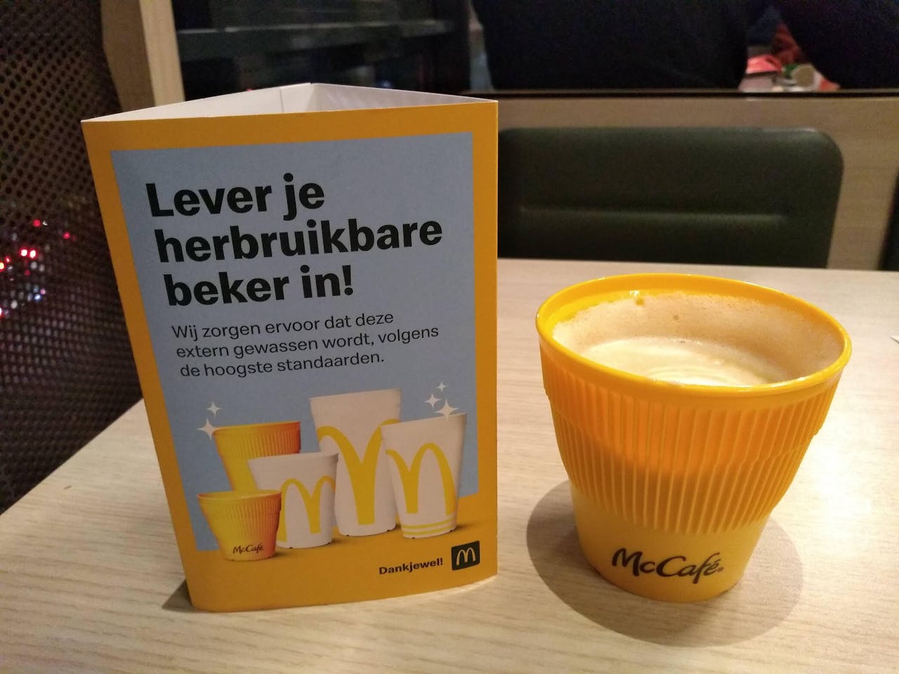 Een gele herbruikbare plastic beker met koffie van McDonald's met daarnaast een bordje met de tekst 'Lever je herbruikbare beker in!'.