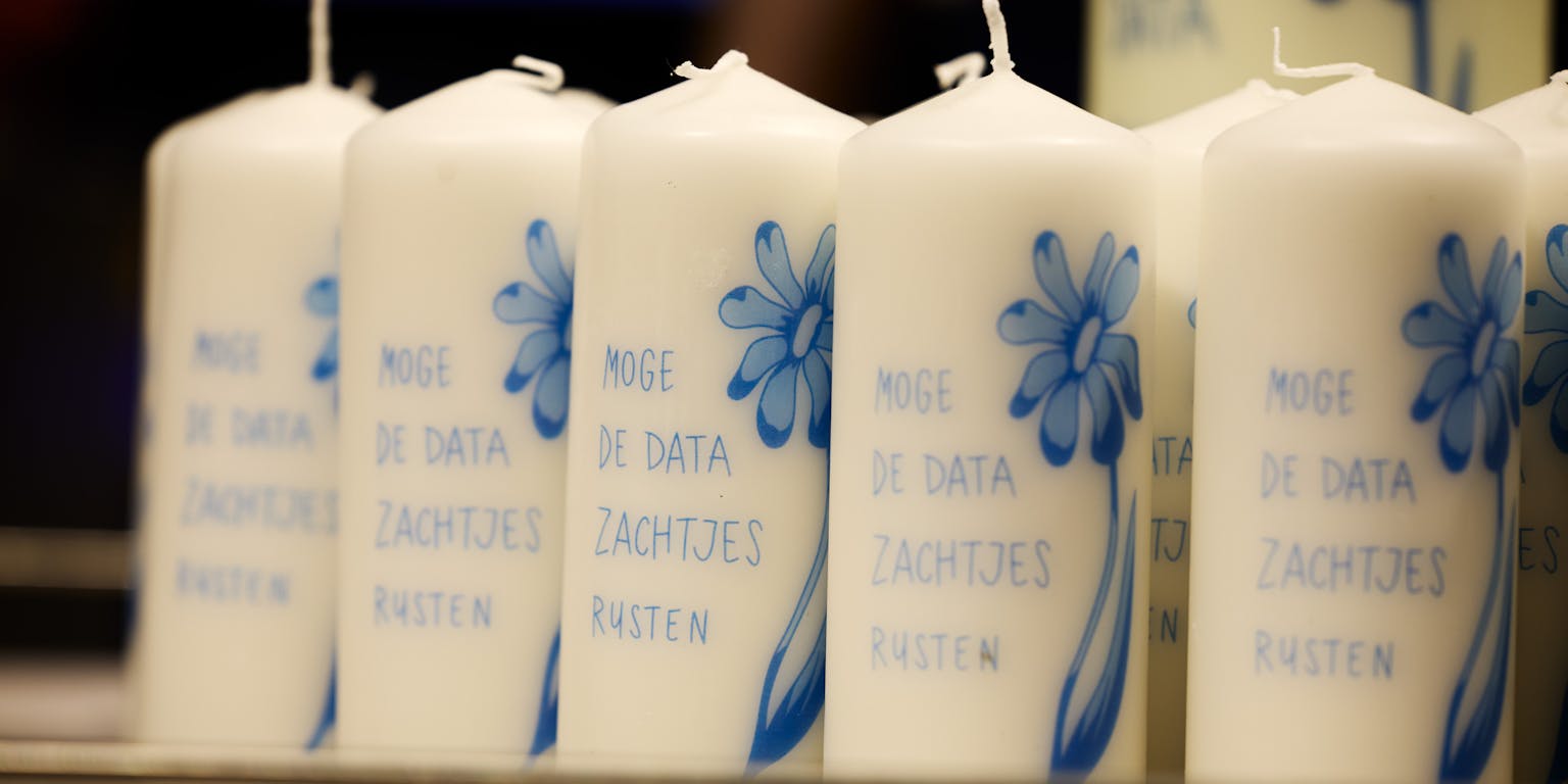 Close-up van witte kaarsen met daarop een blauwe bloem en de blauwe tekst 'Moge de data zachtjes rusten'.