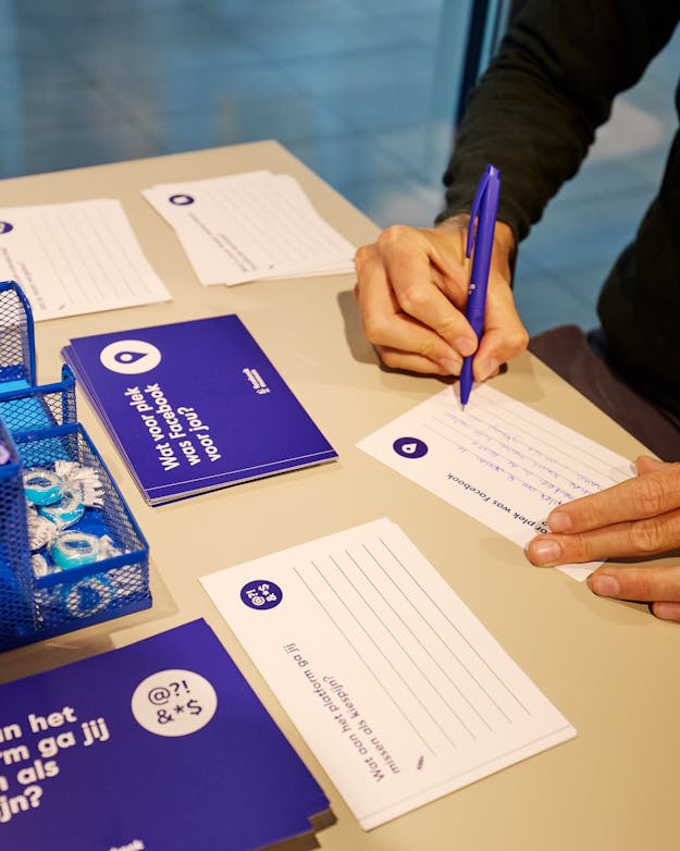 Een bezoeker van het Facebook Museum schrijft een herinnering op een kaartje. Op een tafel liggen kaarten, blauwe voorkant, witte achterkant met ruimte om te schrijven. Een linkerhand houdt een kaartje vast, de rechterhand beschrijft met een blauwe pen het kaartje. Ook staat er een blauwe pennenbak met blauwe pennen op tafel.