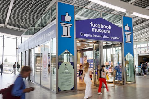 Het tijdelijke Facebook Museum, tussen spoor 19 en 20 op Utrecht Centraal. In een hal met grijs dak staat een grote kas met blauwe rand langs het dak. Mensen lopen voorbij.