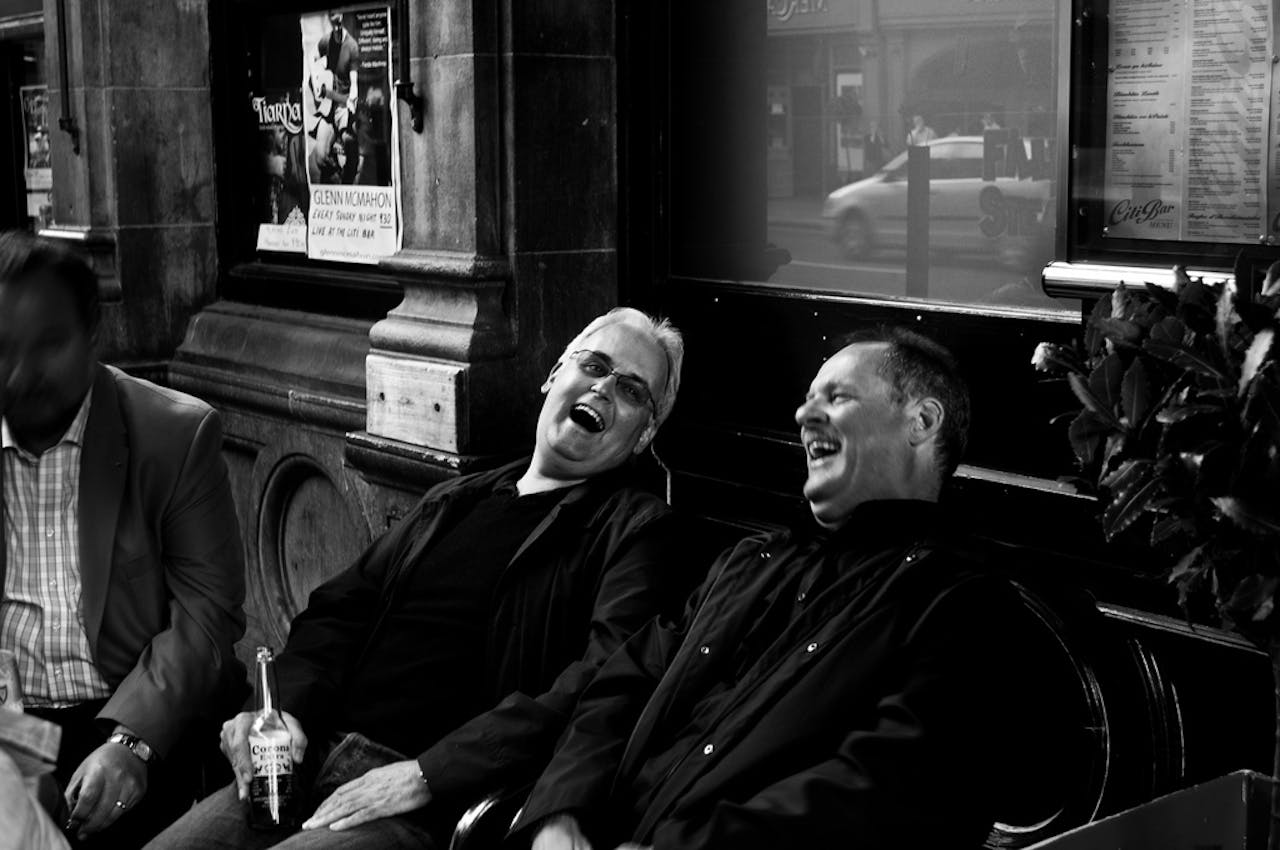 Drie lachende mannen zitten op een bankje voor een gebouw. De afbeelding is zwart-wit.