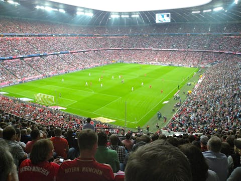 De Allianz Arena in München biedt plaats aan ruim 75.000 toeschouwers. Voetbalveld in een stadion met tribunes in drie lagen om het veld heen.