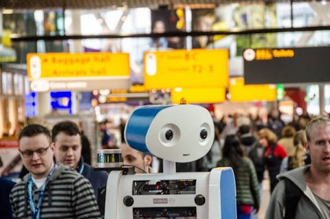 Robot Spencer bracht passagiers op Schiphol naar hun gate van vertrek. Een witte mensachtige robot kijkt om zich heen in een drukke hal van een vliegveld.
