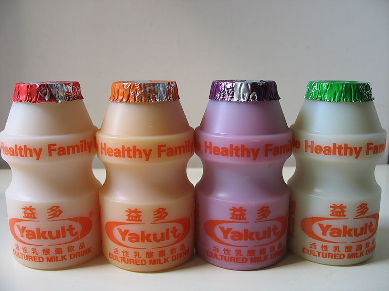 Vier flesjes van Yakult op een rij in vier verschillende smaken. Op de flesjes staat de tekst 'gezonde familie'.