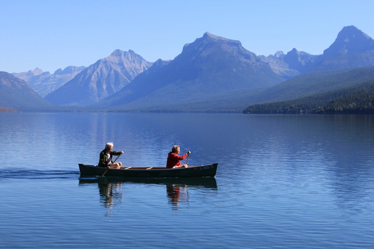 Twee mensen in een kano op open water. Er zijn bergen te zien op de achtergrond.