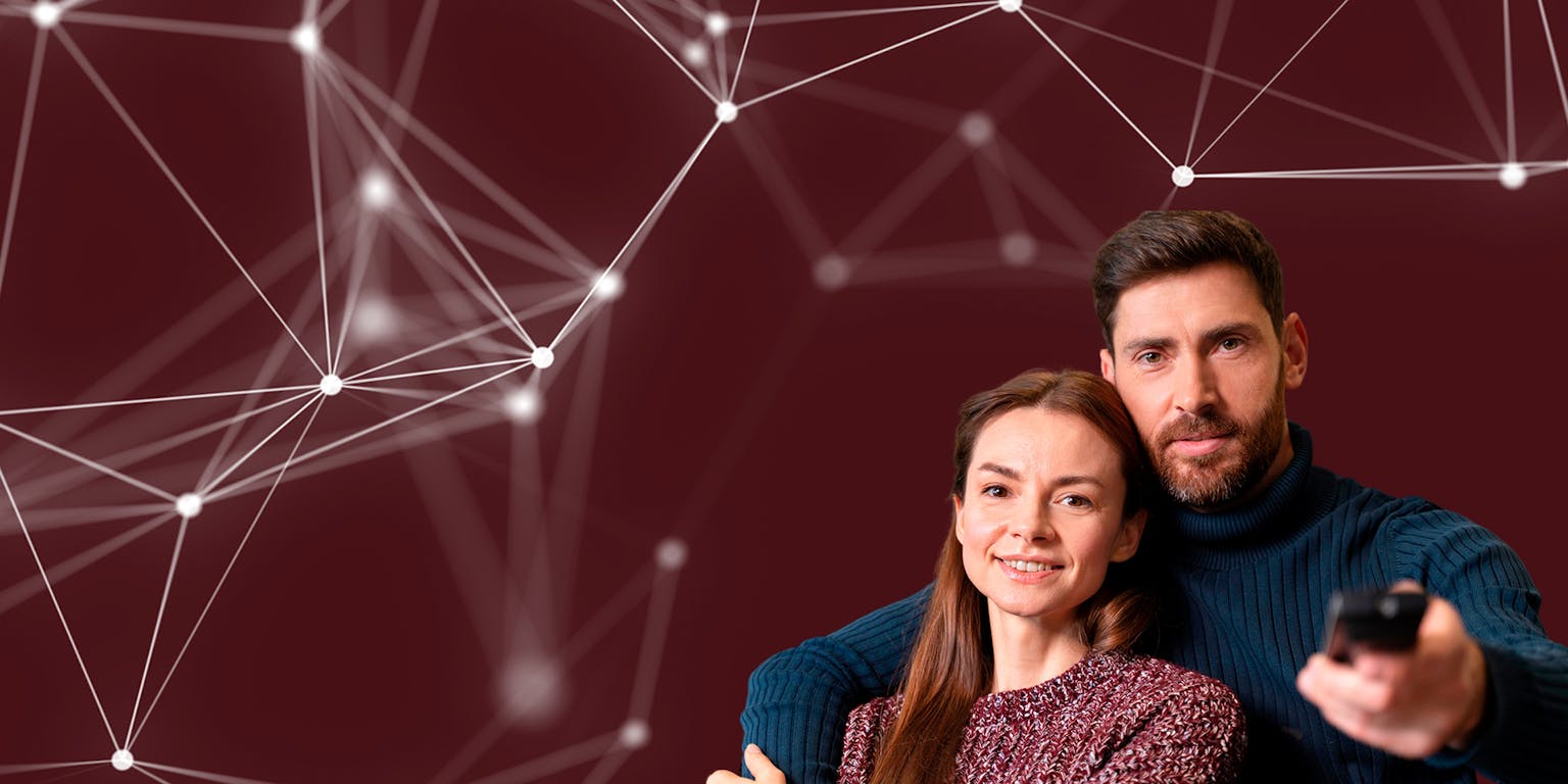 Een man en een vrouw leunen tegen elkaar aan. De man heeft een afstandsbediening in zijn linkerhand en wijst deze richting de camera.