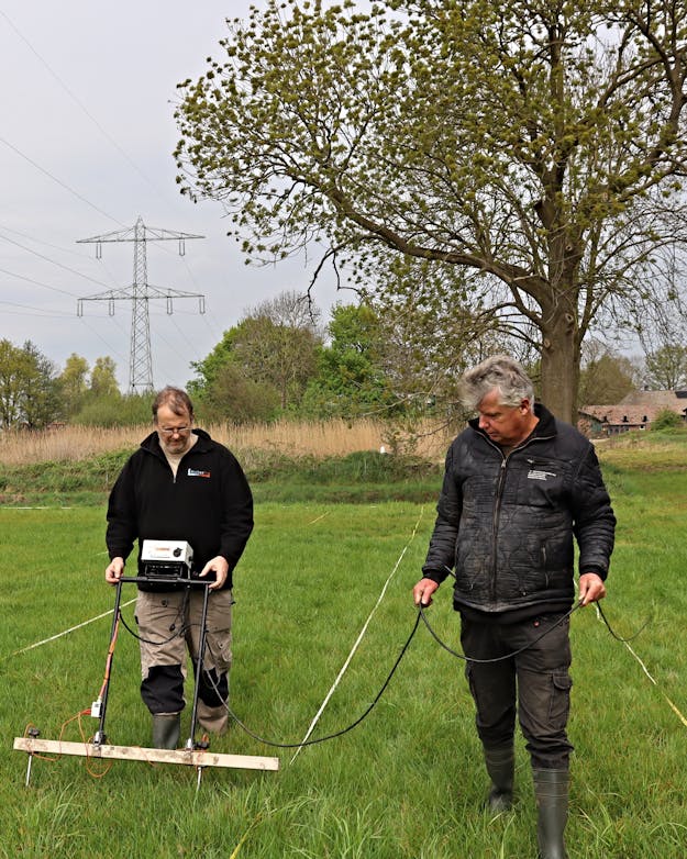 Twee mannen die een archeologische weerstandsmeter in de grond prikken. Onderaan de meter hangt een horizontale balk met daaraan twee punten. Twee mannen die een archeologische weerstandsmeter in de grond prikken. Onderaan de meter hangt een horizontale balk met daaraan twee punten.