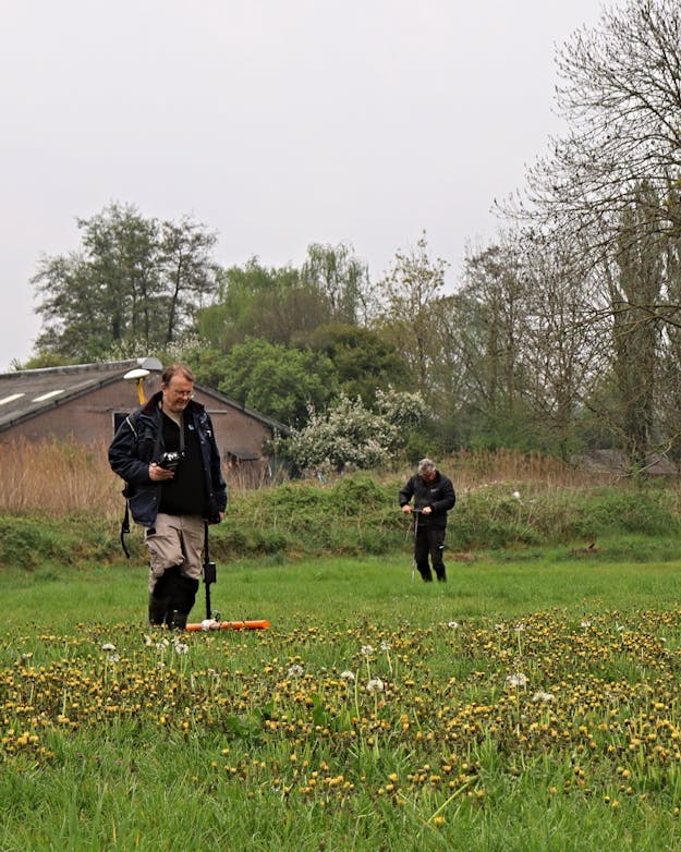 Twee mannen in een grasveld. De ene man heeft een apparaat met een oranje buis vast en de ander steekt een buis in de grond. Twee mannen in een grasveld. De ene man heeft een apparaat met een oranje buis vast en de ander steekt een buis in de grond.