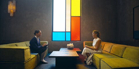 ‘Aspirant-moeder’ Mia (rechts) en assessor Virginia (links), tegenover elkaar in ‘The Assessment’. Scene uit de film The Assessment, waarin Mia (Elizabeth Olsen, rechts) en Virginia (Alicia Vikander, links) tegenover elkaar op de bank zitten. Er staat een lage koffietafel tussen hen in