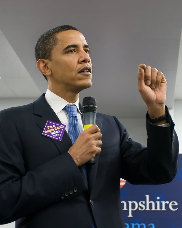 Obama houdt in zijn ene hand een microfoon, en maakt met zijn andere hand een knijpgebaar. Obama houdt in zijn ene hand een microfoon, en maakt met zijn andere hand een knijpgebaar.