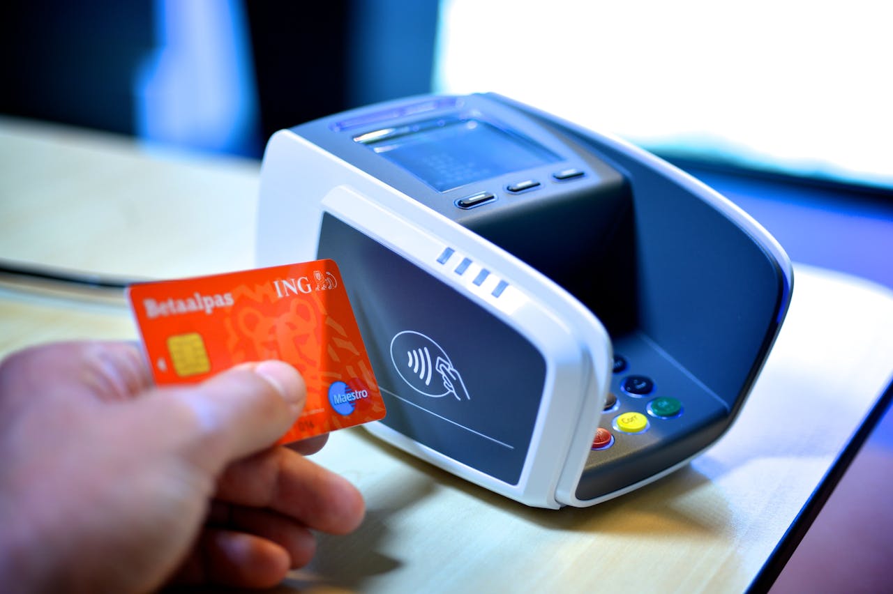 Een hand houdt een oranje bankpas bij een grijs pinautomaat. Op de zijkant is het logo voor contactloos betalen te zien.