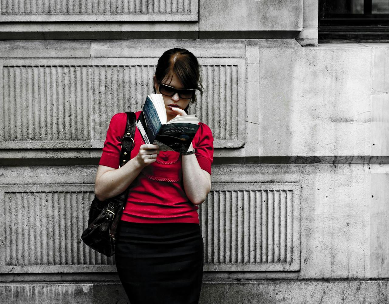 Een persoon met een zonnebril op en een rode trui aan, leunt tegen een muur en leest een boek.