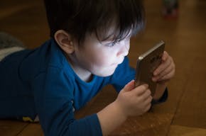 Een jongetje ligt op zijn buik en kijkt op een smartphone. Zijn gezicht is verlicht door het scherm.