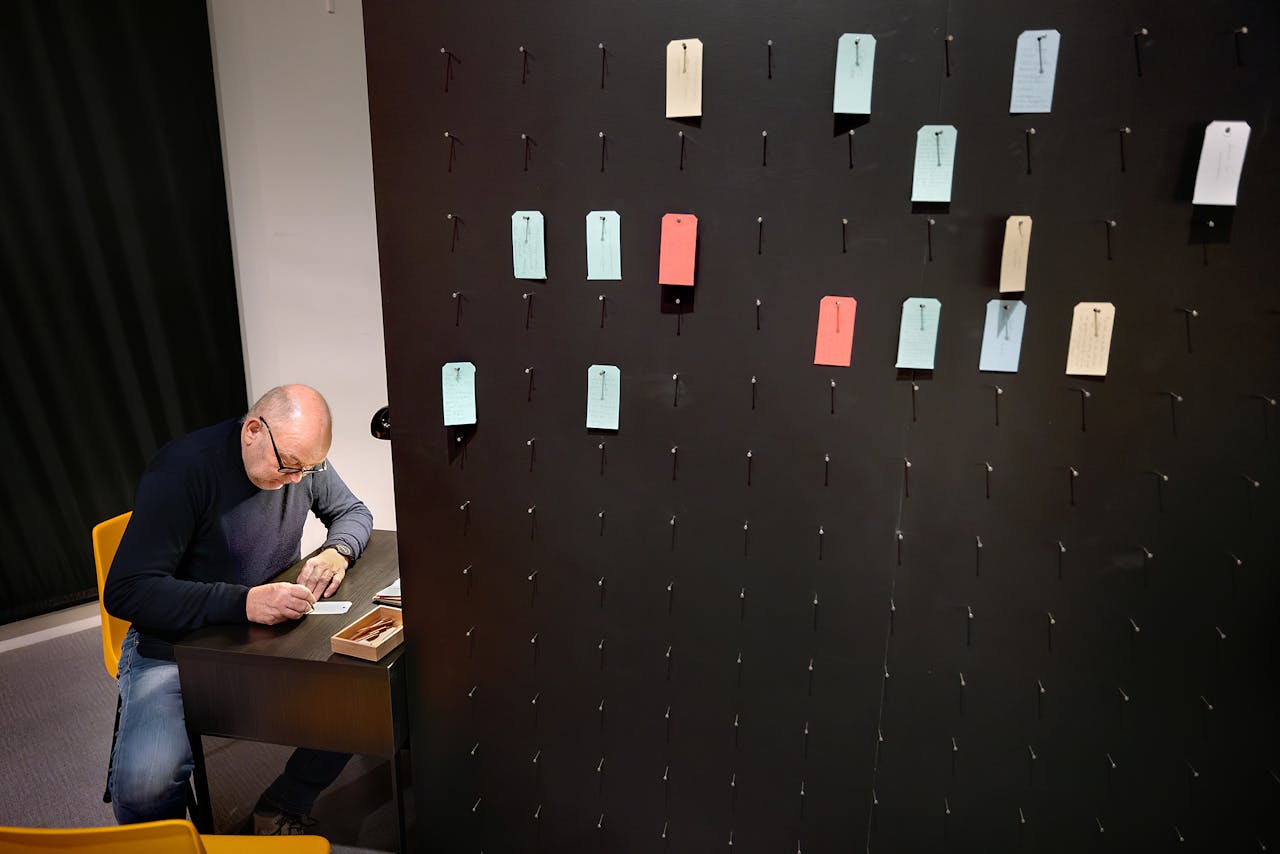 Een bezoeker van de tentoonstelling schrijft een briefje aan een bureau achter een wand waar nog meer briefjes ophangen.
