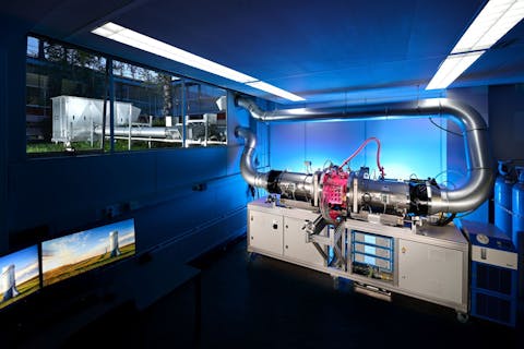 Het prototype van de CO2-afvanginstallatie van Carbyon. Een grijs apparaat in een donkere, blauw verlichte ruimte. Een buis loopt van het apparaat door de muur naar buiten. Door het raam is de buitenunit van het apparaat te zien.