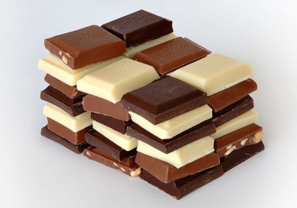 Een stapel chocoladeblokjes in wit, melk en puur gemixt door elkaar op een witte achtergrond.