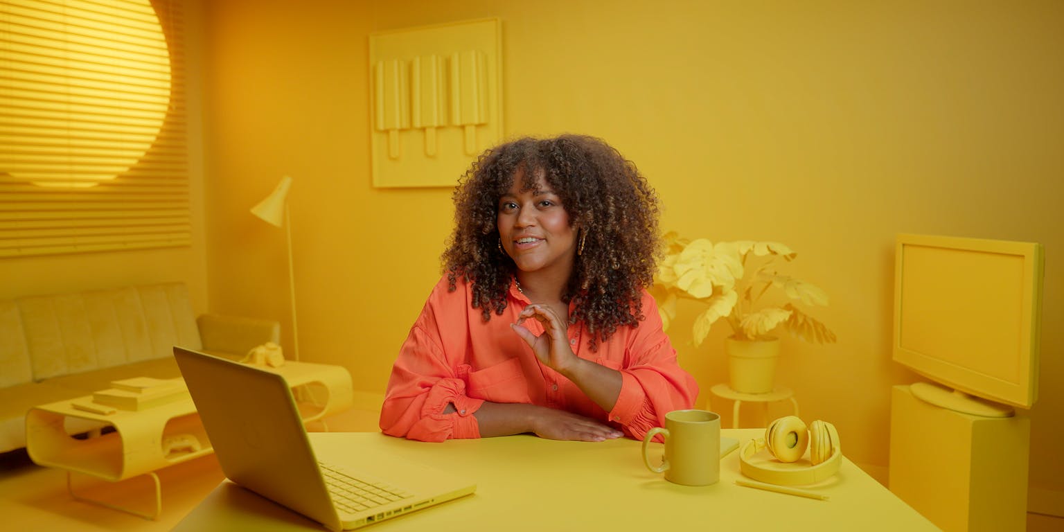 Presentatrice Quinty Misiedjan, een jongedame met lichtgetinte huidskleur en fijne, donkere krullen, spreekt tegen de camera. Ze draagt een oranje blouse en zit aan tafel in een kamer waar alles geel is.