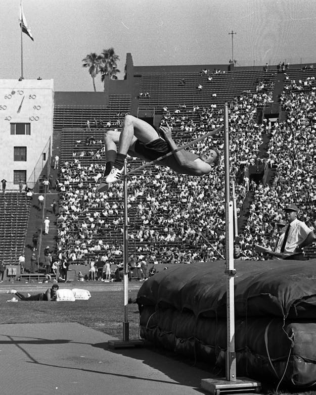 Hoogspringer Dick Fosbury tijdens de Olympische Spelen in 1968. Zwartwitfoto van een hoogspringer die achterwaarts over de lat heen springt. Op de achtergrond een tribune vol toeschouwers.