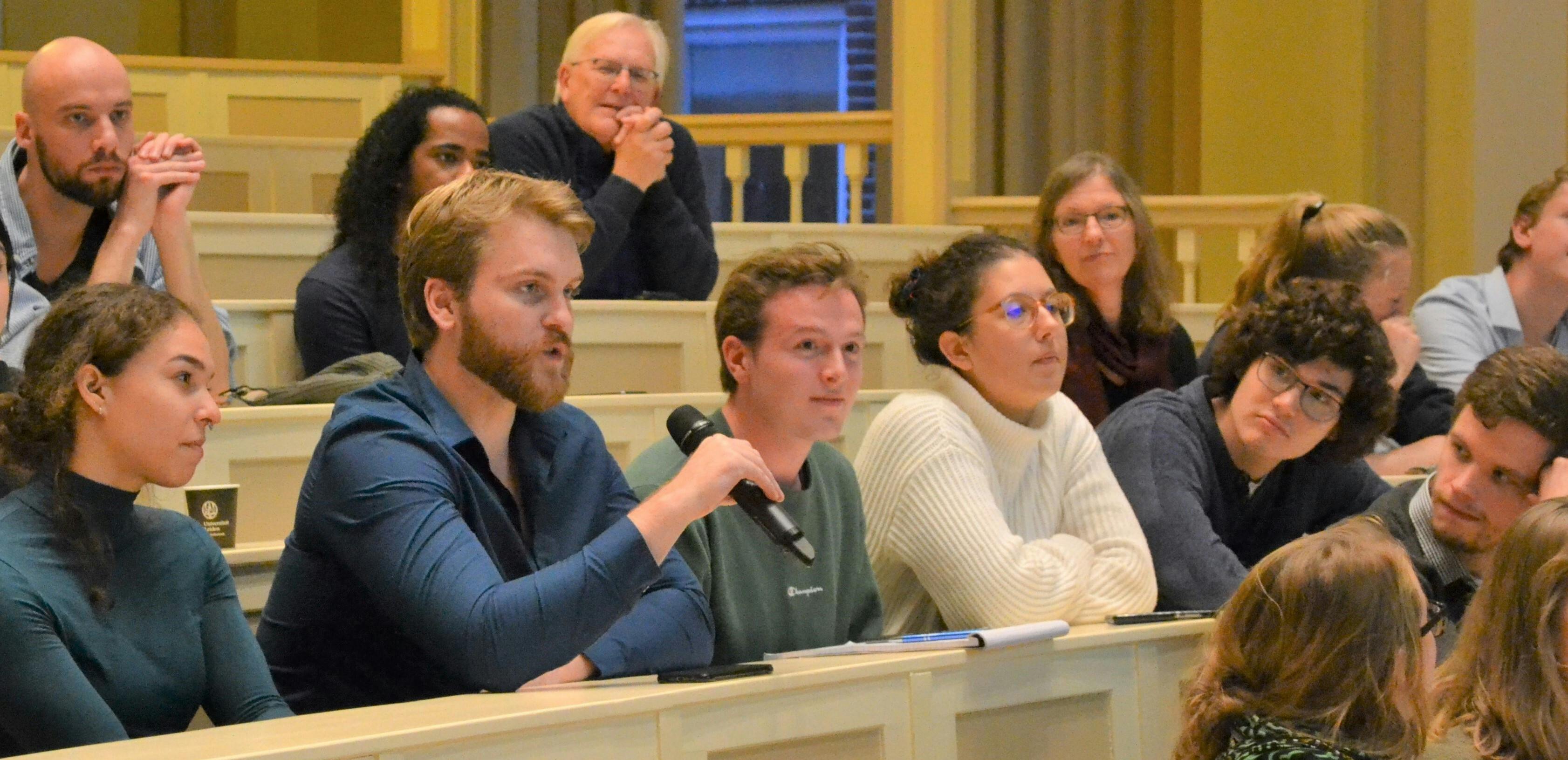 Een groep mensen zit in een collegezaal. Een persoon heeft een microfoon in zijn handen.