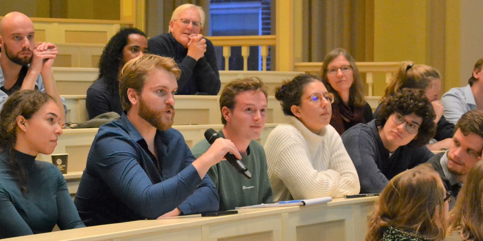 Een groep mensen zit in een collegezaal. Een persoon heeft een microfoon in zijn handen.