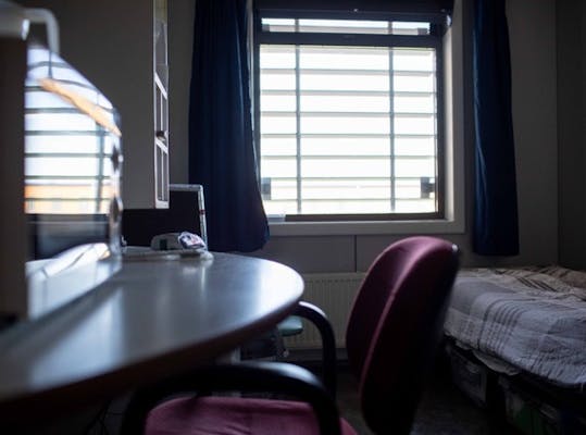 Cel in een gevangenis met een raam met tralies ervoor, een bed en een bureautje met bureaustoel en magnetron