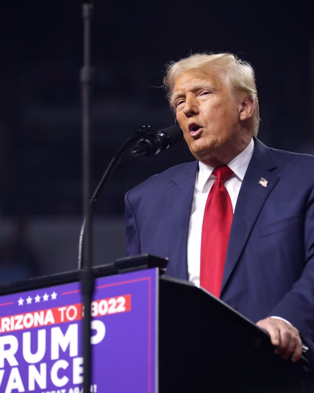 De Amerikaanse presidentskandidaat Donald Trump tijdens de verkiezingscampagne in 2024. De Amerikaanse presidentskandidaat Donald Trump staat achter een spreekgestoelte met microfoon.