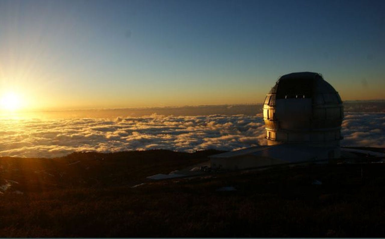 Een foto van de Gran Tecan telescoop op La Palma boven de wolken. De zon is duidelijk zichtbaar.